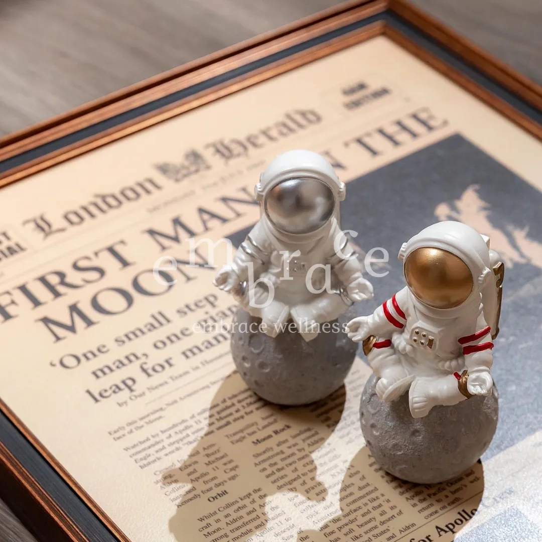Pair of meditating astronaut ornaments displayed with vintage moon‑landing newspaper, space‑themed home decor.