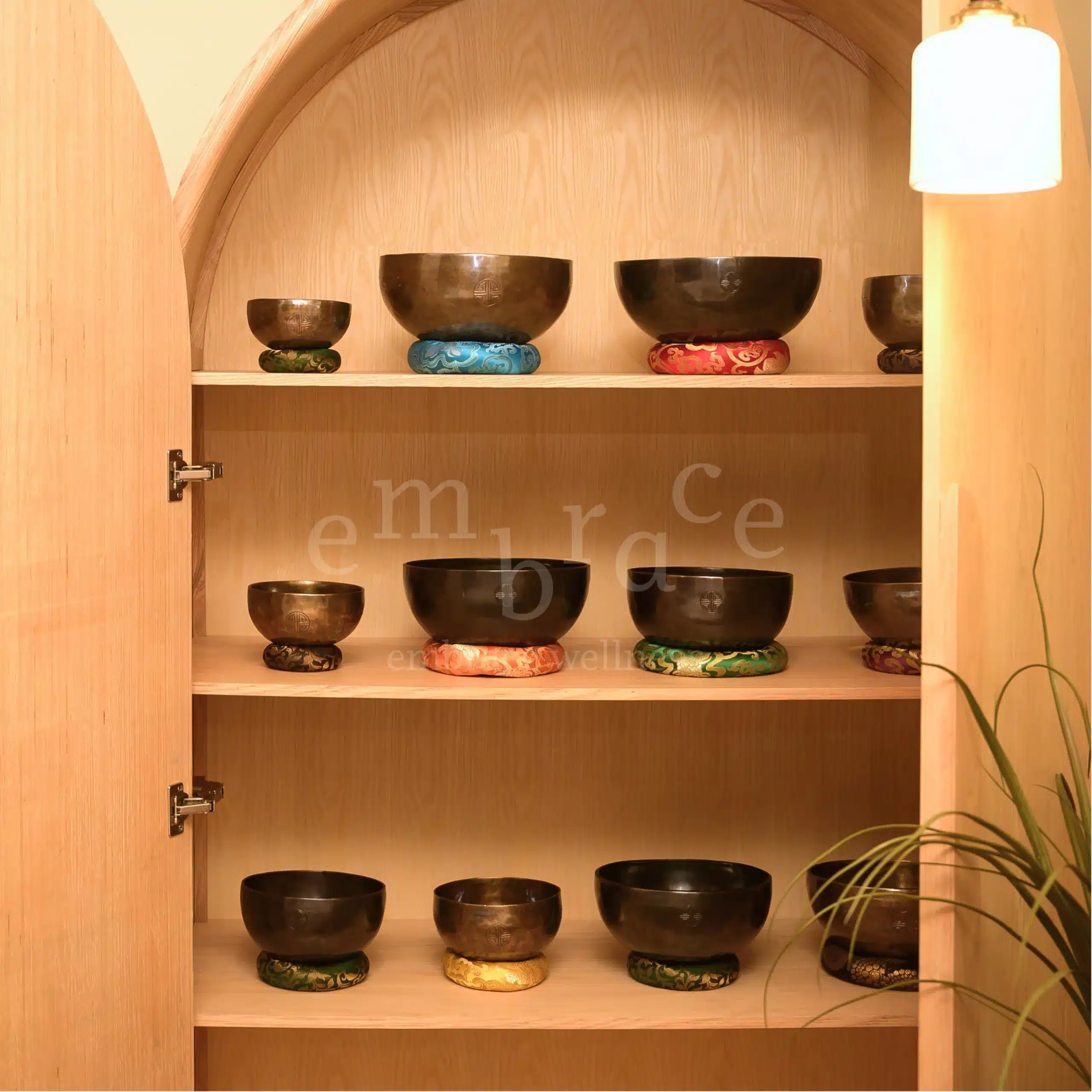 A beautifully crafted arched wooden cabinet open to display a curated collection of Tibetan singing bowls by professional and experienced sound healer Manlee from Embrace Wellness based in Newbury Berkshire, on colourful cushioned rings — the sound healing instrument collection at Embrace Wellness, Newbury, Berkshire