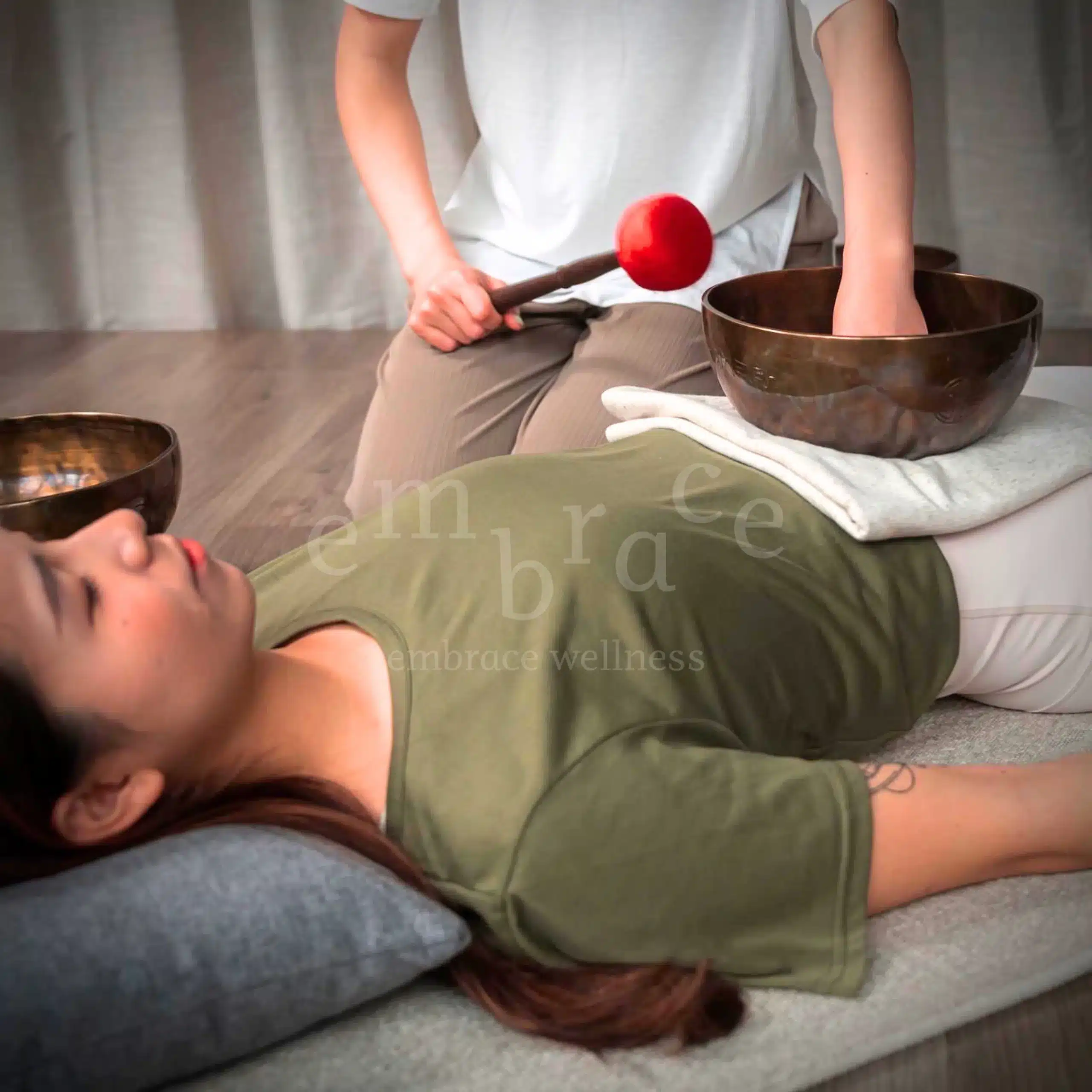 A client lying down in deep relaxation during a Tibetan singing bowl sound healing session at Embrace Wellness, with a large antique bowl placed on the body and a red-tipped mallet being held above it by the sound healer