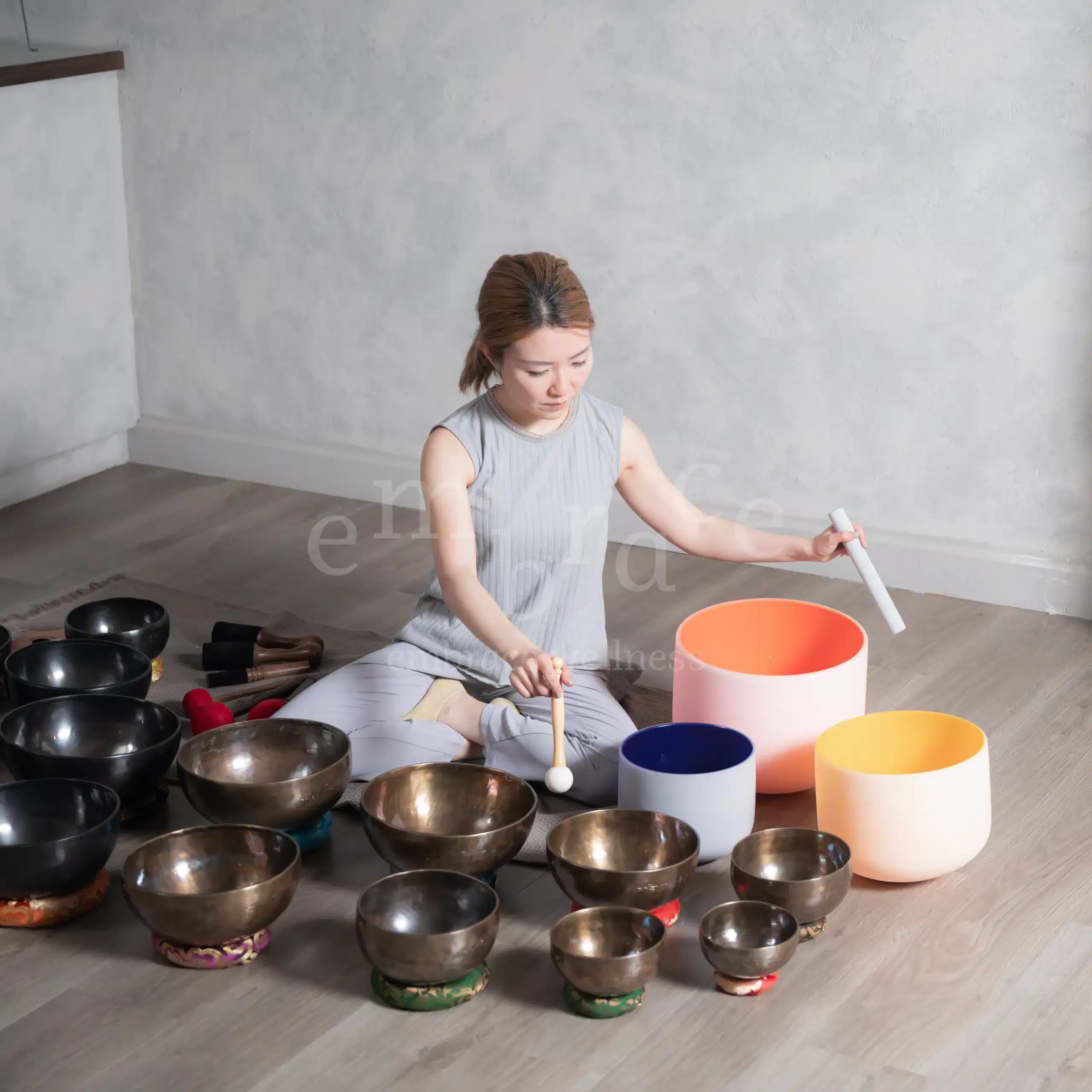 Professional and experienced sound healer Manlee W at Embrace Wellness Newbury sitting cross-legged surrounded by a full collection of antique Tibetan singing bowls and colourful frosted crystal singing bowls, playing two mallets simultaneously