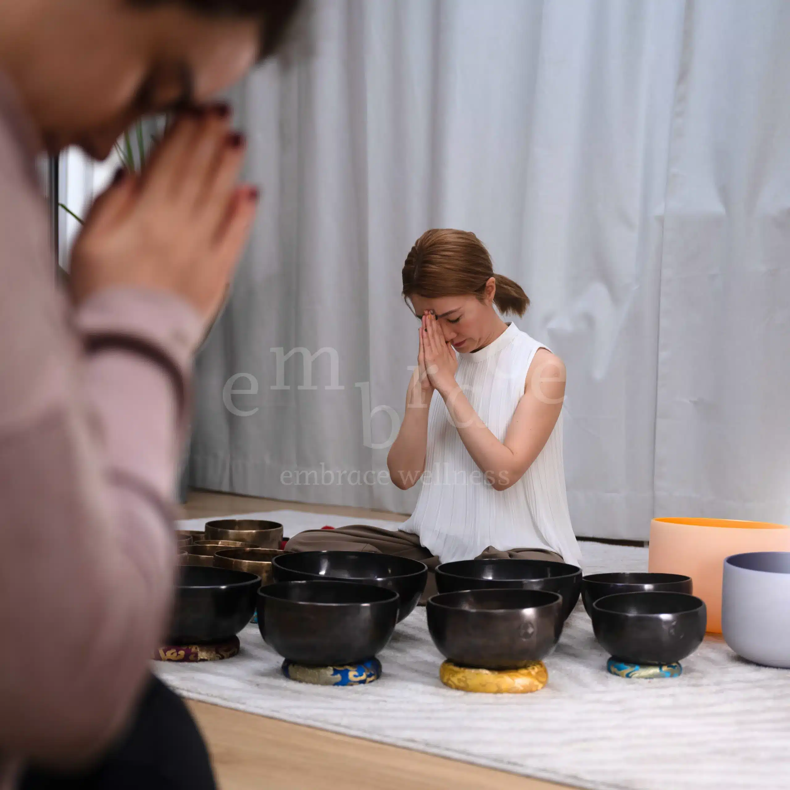 Professional and experienced sound healer Manlee W sitting in prayer position behind a ceremonial arrangement of Tibetan singing bowls, with a client mirroring the gesture in the foreground, marking the opening of a sound healing session