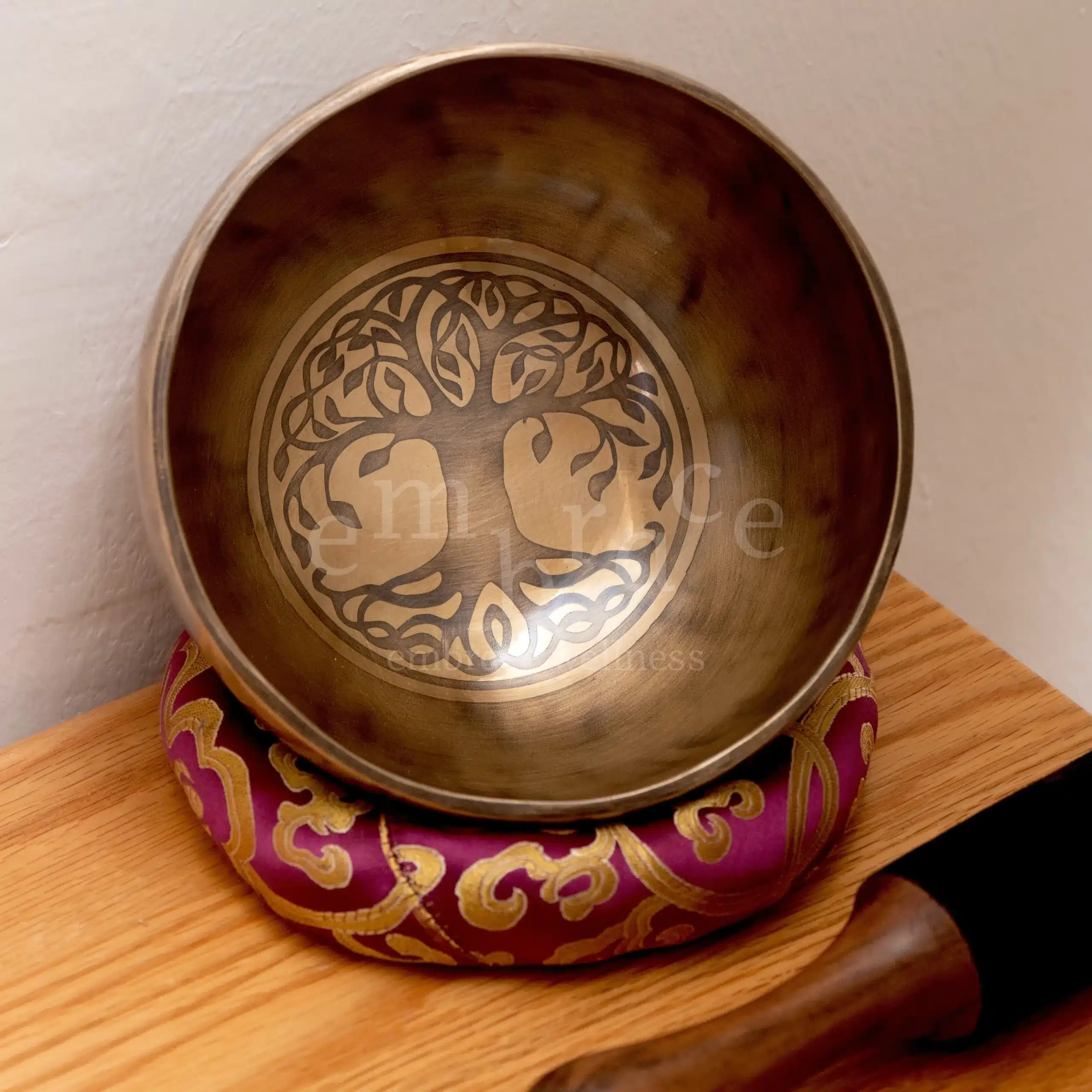 Close-up of an antique Tibetan singing bowl with an engraved Tree of Life symbol inside, resting on a deep purple and gold embroidered cushion on a wooden surface at Embrace Wellness Newbury