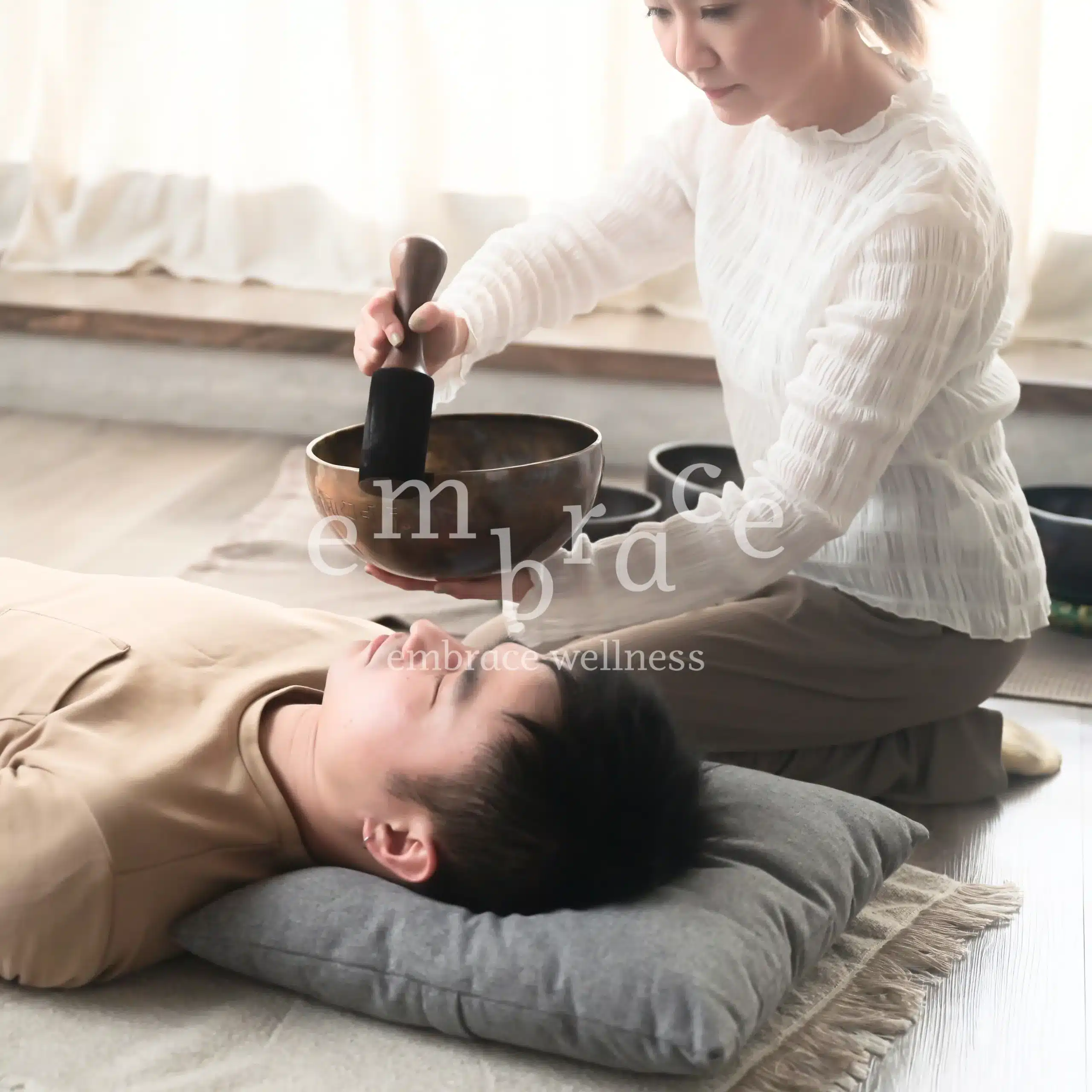 Professional and experienced sound healer Manlee W holding a Tibetan singing bowl close to a client lying peacefully on a cushion during a sound healing session at Embrace Wellness, Newbury, West Berkshire.