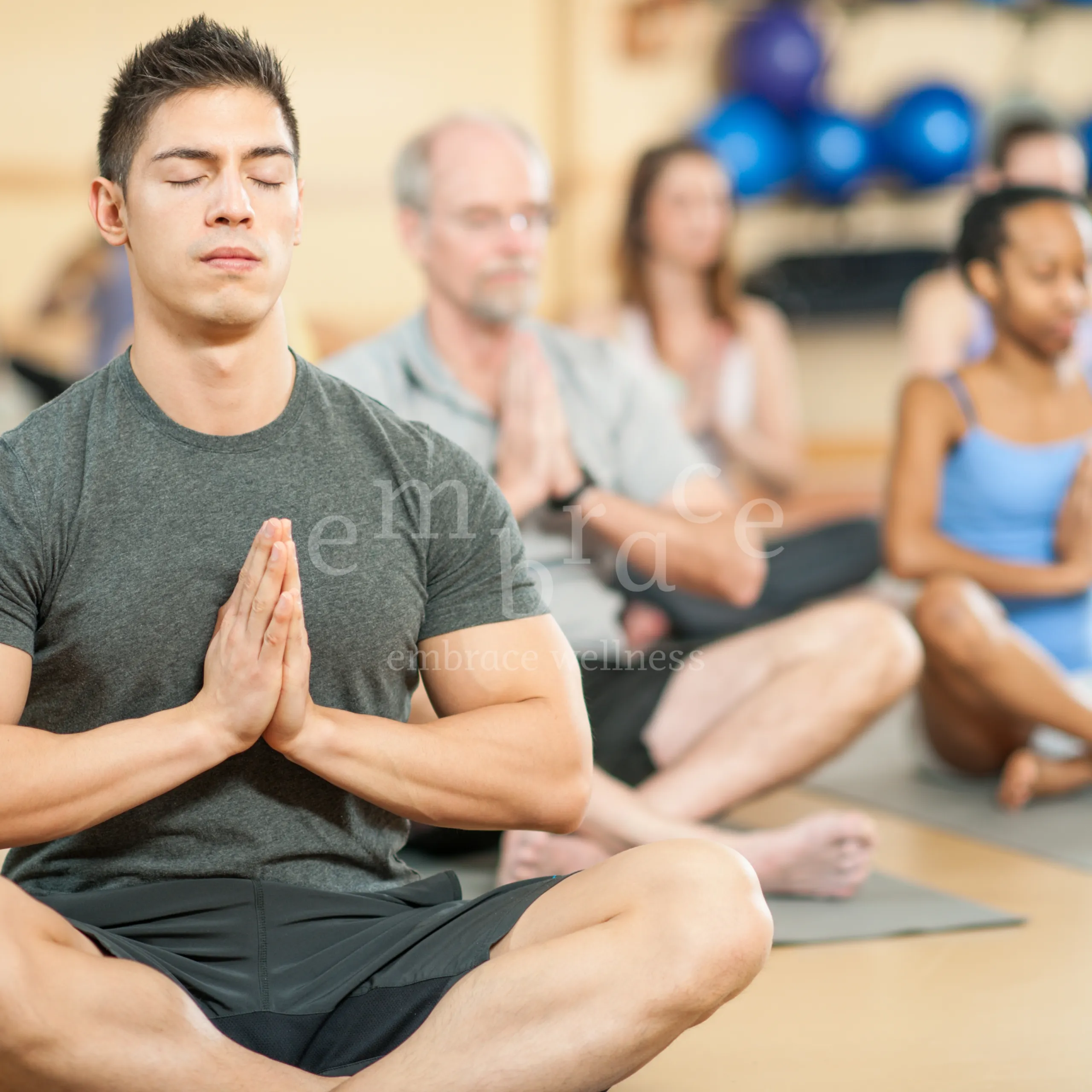 A diverse group of men and women of all ages seated in cross-legged meditation with prayer hands (Anjali mudra) during a beginner yoga class in Newbury with Embrace Wellness by Manlee W